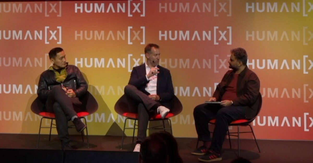 Three men seated on stage discussing at Human[X] event, with branded backdrop and one holding a notepad.